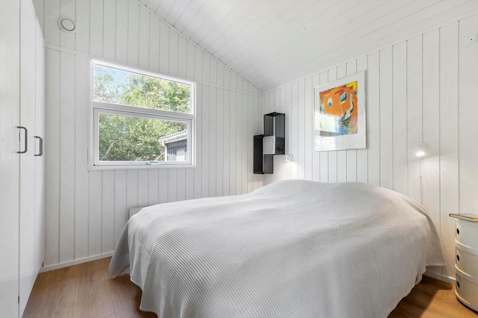 Schlafzimmer mit weißer Holzwand, Bett, Fenster und Wandbild.