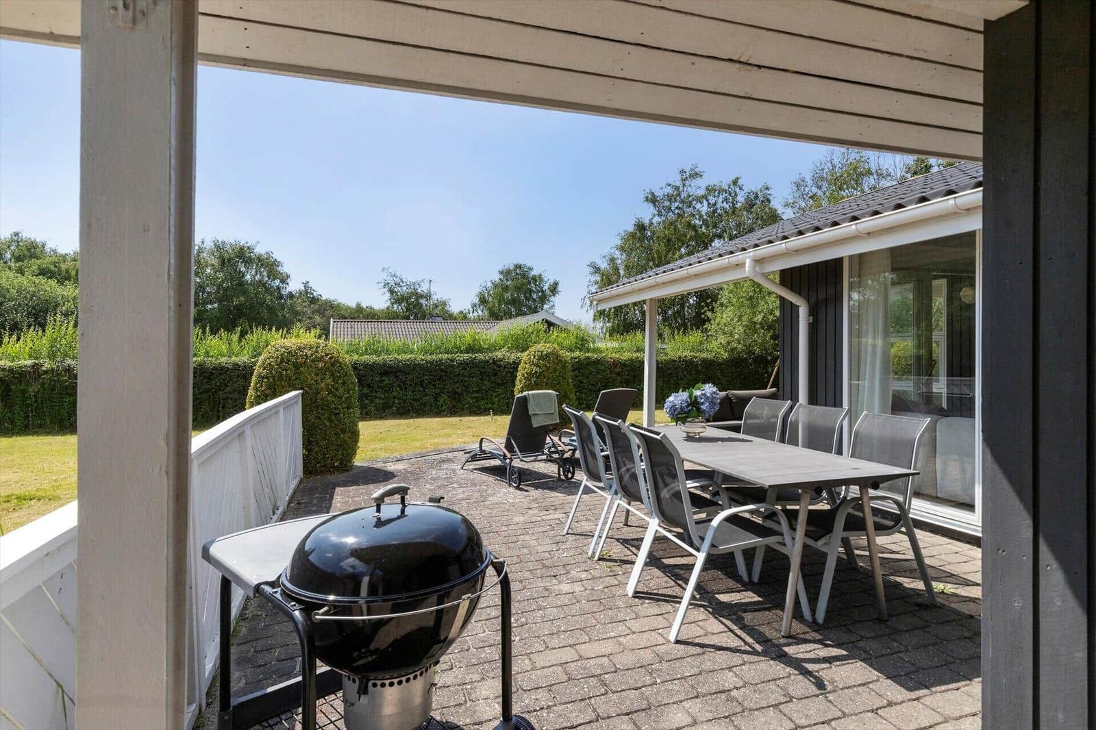 Terrasse mit Grill, Tisch und Stühlen. Hintergrund: Garten mit Hecke und Bäumen.