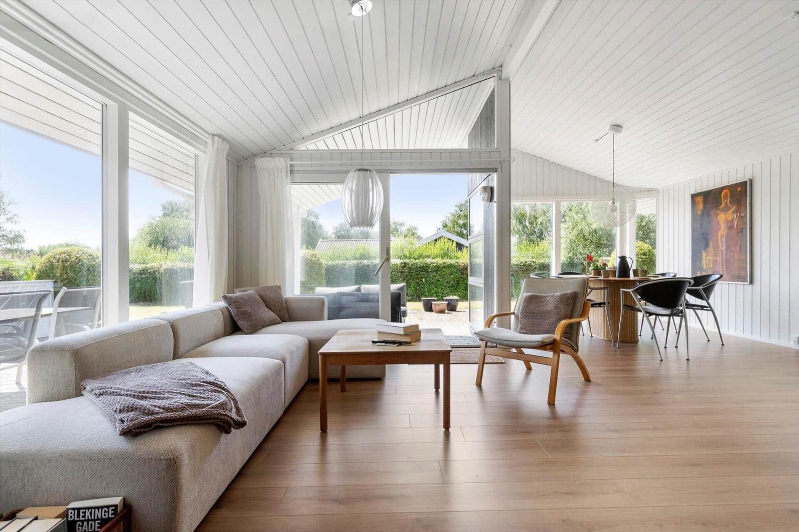 Wohn- und Essbereich mit Sofa, Stuhl und Tisch. Große Fenster und Tür zu Garten.