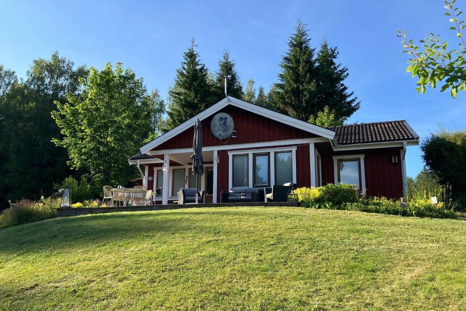 Rotes Holzhaus mit Terrasse, Garten und Bäumen im Hintergrund.
