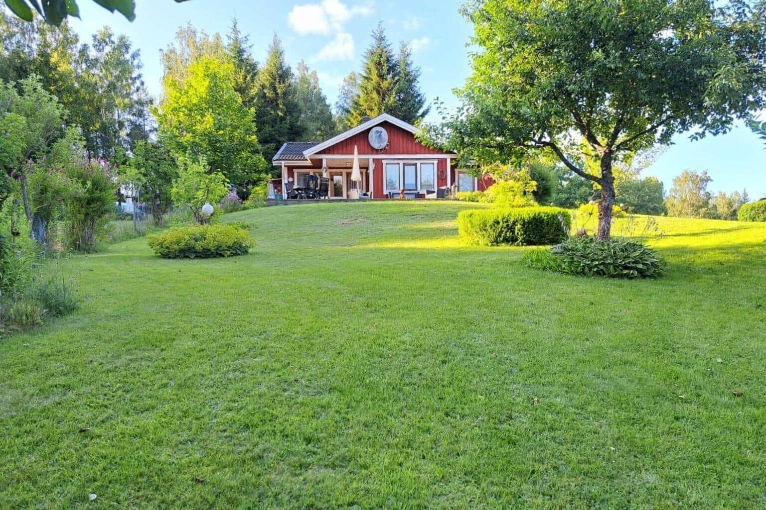 Rotes Haus mit Terrasse und Garten. Umgeben von Bäumen und Grasflächen.