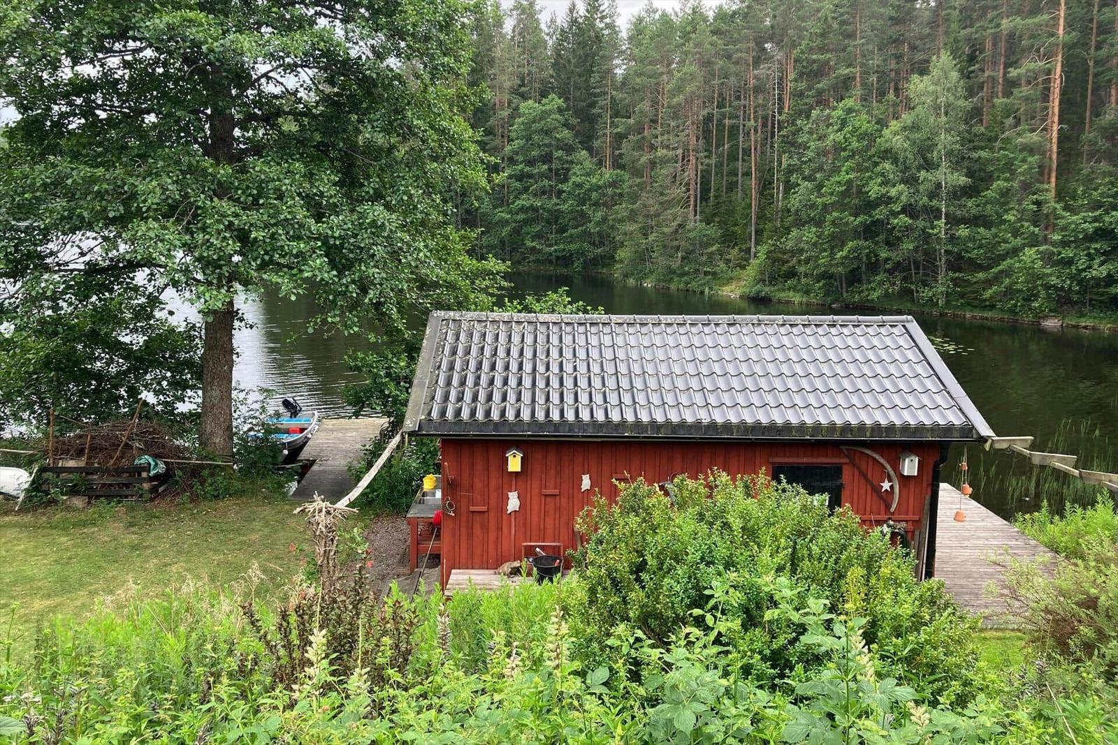 Rotes Holzhaus am See mit Boot und Holzsteg. Umgeben von Wald und Grünflächen.