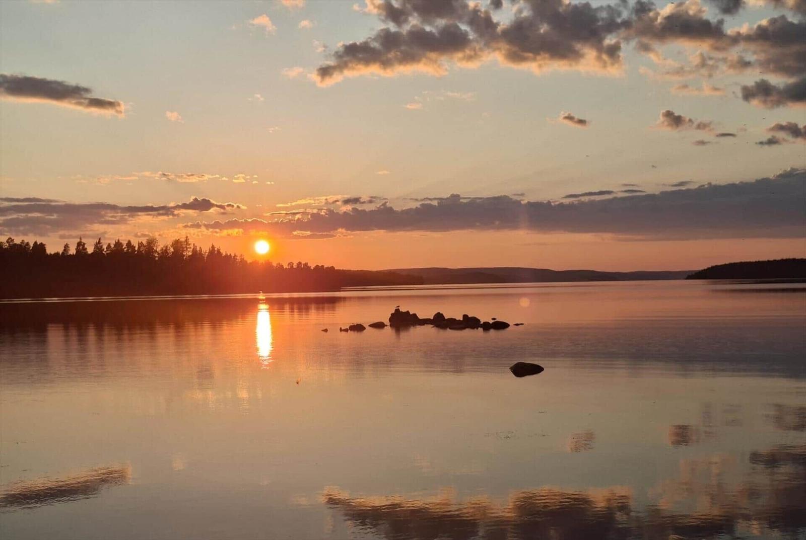 Sonnenuntergang über ruhigem See mit Reflexion und Felsen im Vordergrund.