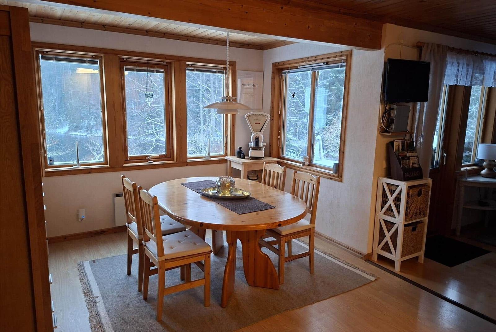 Esszimmer mit Holztisch, Stühlen und Fenstern zum Wald. Fernseher und Schrank sind sichtbar.
