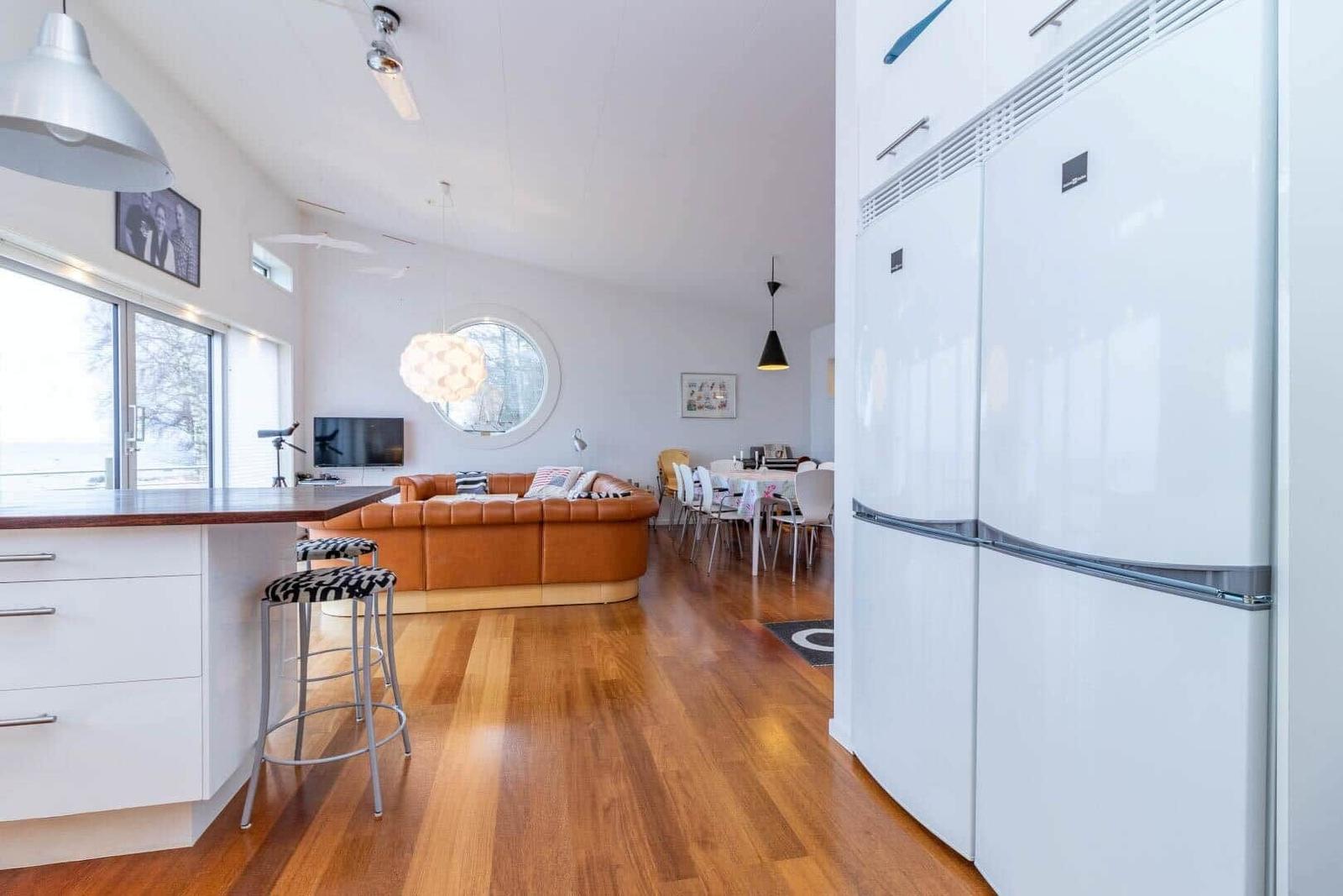 Küche mit weißen Schränken und Kühlschrank. Wohnbereich mit braunem Sofa und runden Fenster.