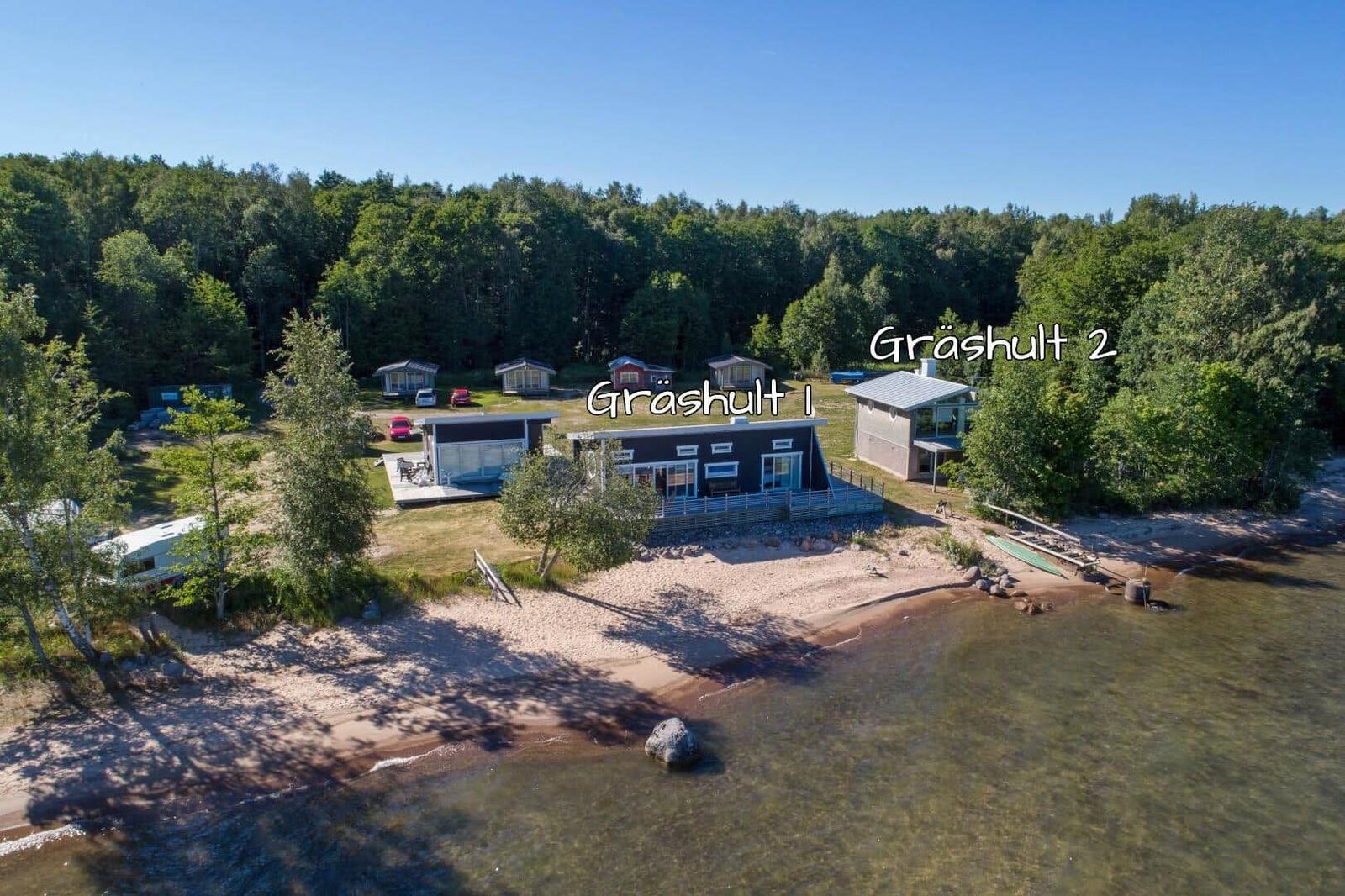 Zwei Häuser am Ufer mit Sandstrand und Wald im Hintergrund.