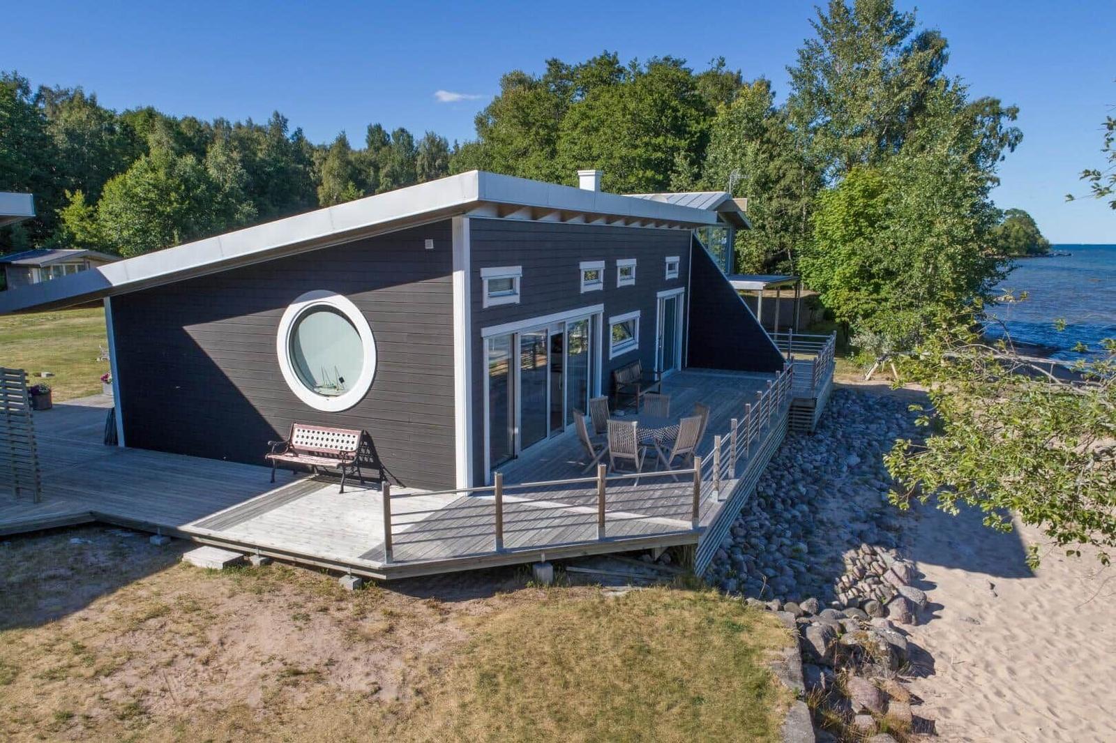 Moderne Strandhütte mit Terrasse, runden Fenster und Blick aufs Wasser.