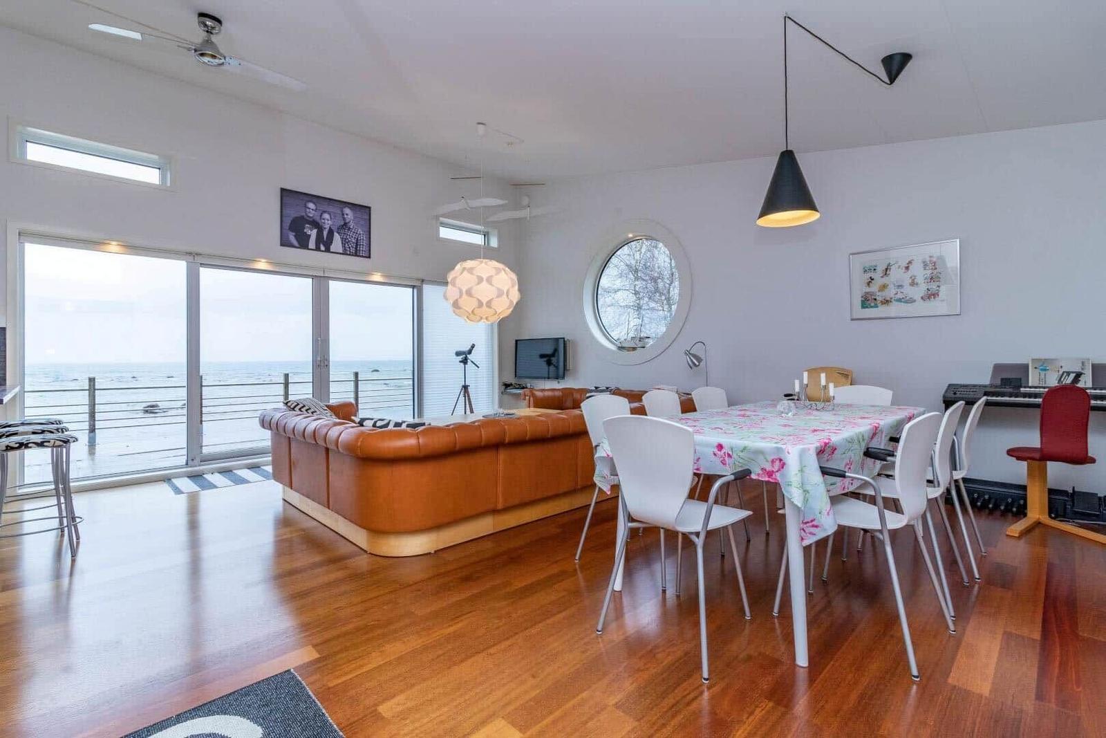 Wohn- und Essbereich mit Blick aufs Meer, brauner Ledersofa, runder Fenster und Esstisch mit Stühlen.