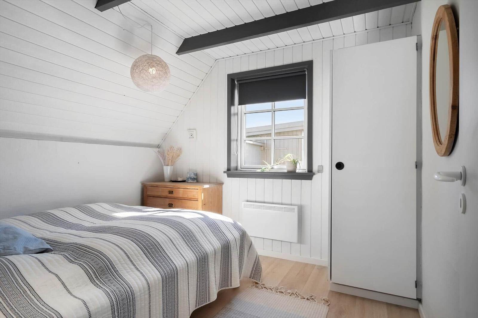 Schlafzimmer mit Bett, Nachttisch und Fenster. Weiße Wände, Holzboden und Deckenbalken.