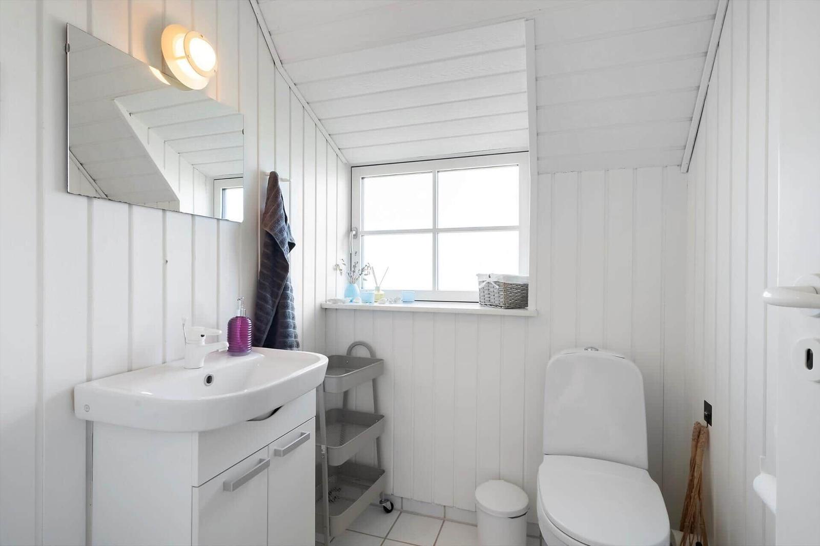 Badezimmer mit Waschbecken, Toilette und Fenster. Weiße Holzwandverkleidung und Spiegel.
