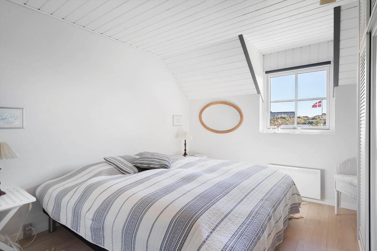 Schlafzimmer mit Bett, Fenster und Holzboden. Decke weiß, Wand mit Spiegel.