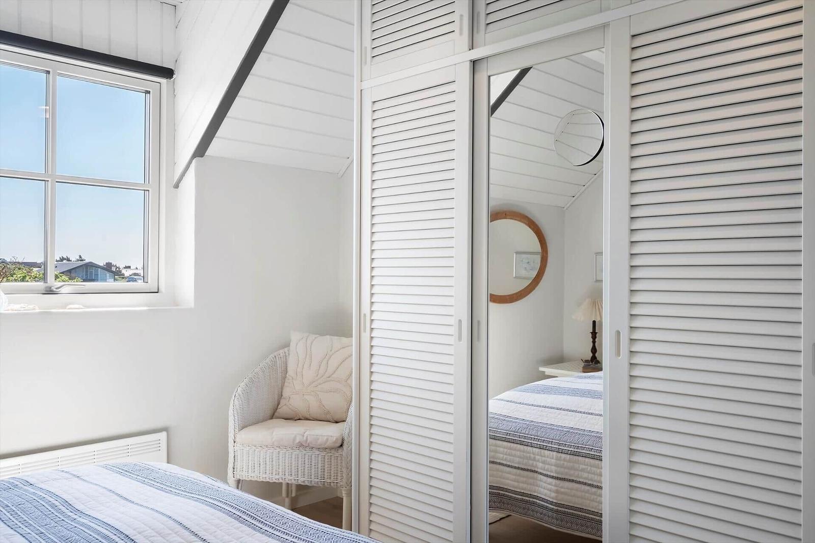 Schlafzimmer mit Fenster, Stuhl und Kleiderschrank mit Spiegeltür.