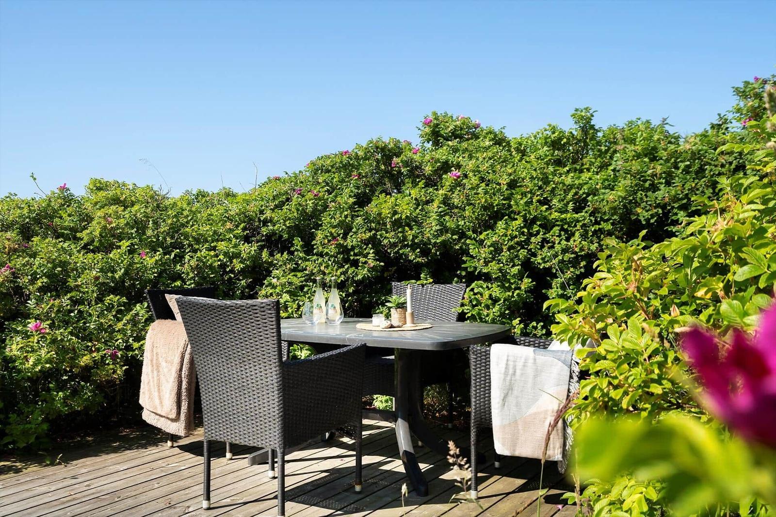 Terrasse mit Tisch und Stühlen, umgeben von grünen Sträuchern und rosa Blüten.