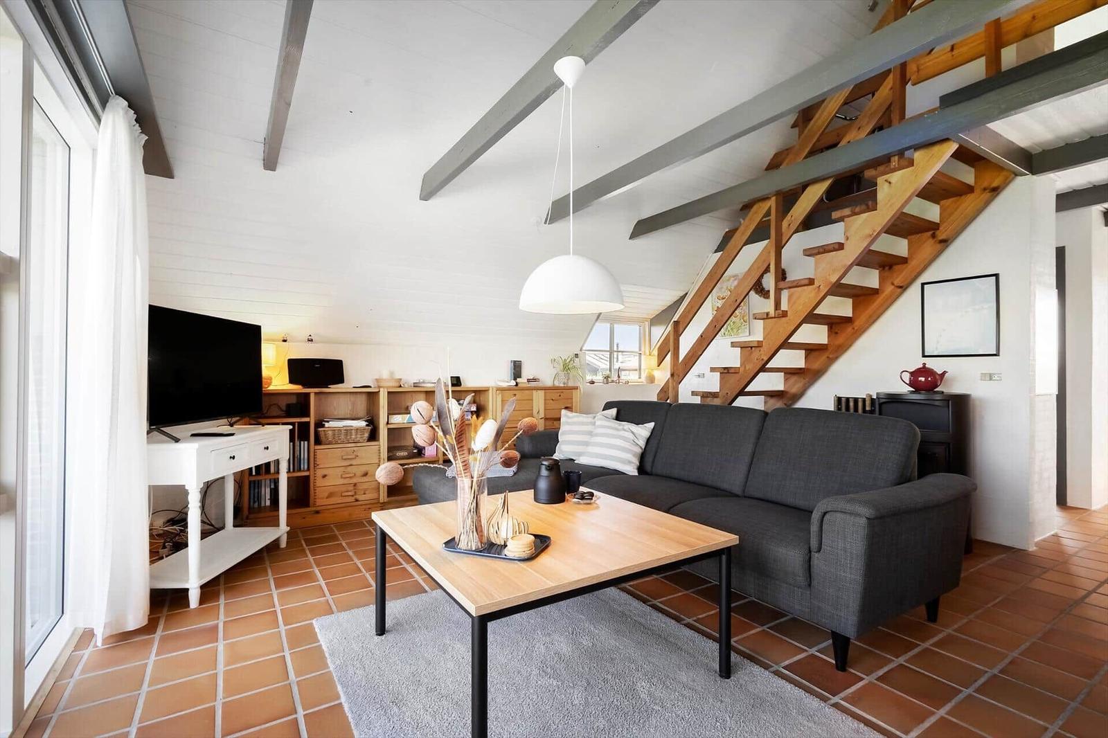 Wohnzimmer mit Sofa, Couchtisch und Holztreppe. Terrakottaboden und weiße Wände.