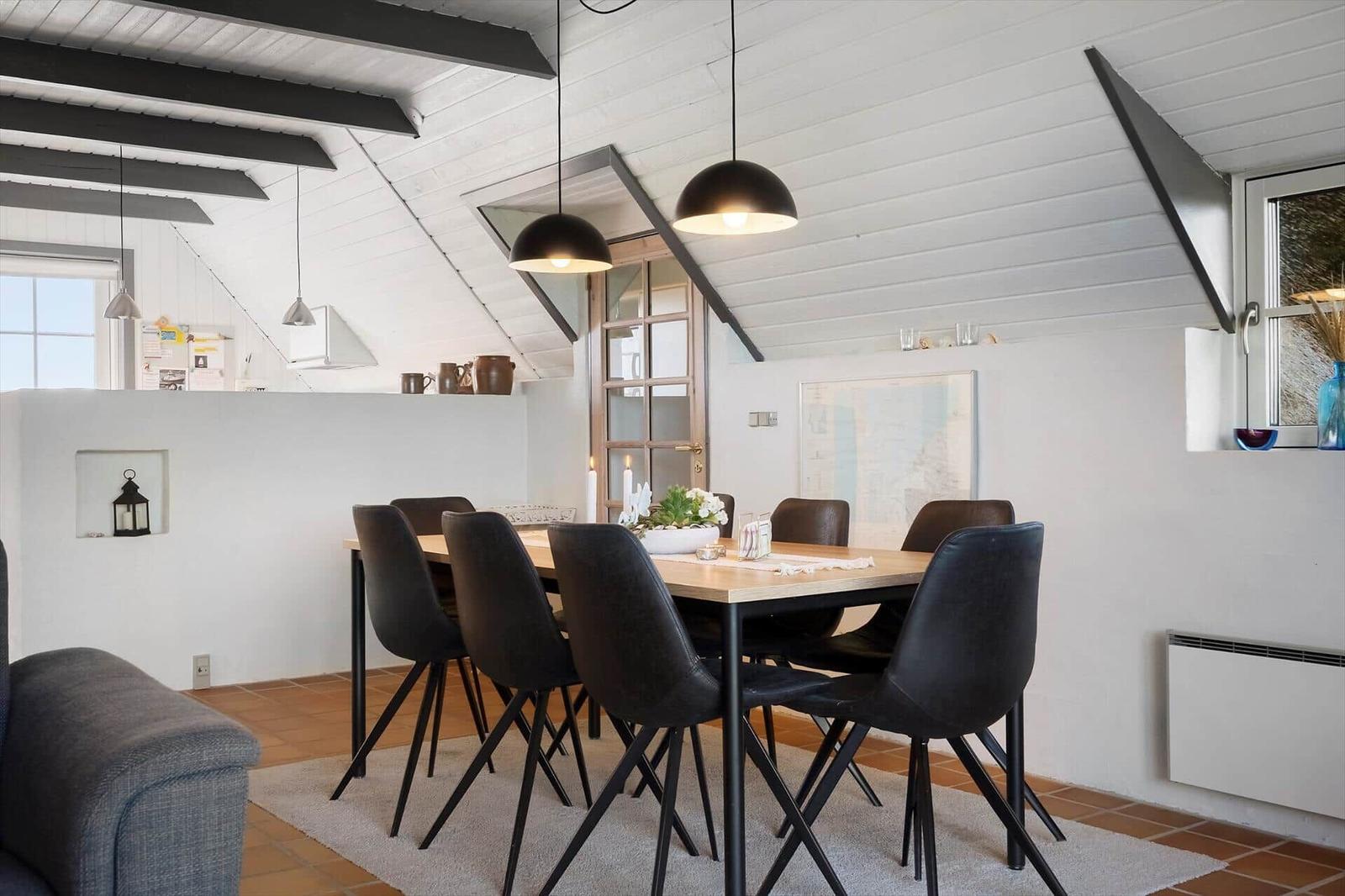 Esszimmer mit Holztisch, schwarzen Stühlen und schwarzem Deckenlicht. Weiße Wände und Holzbalken.