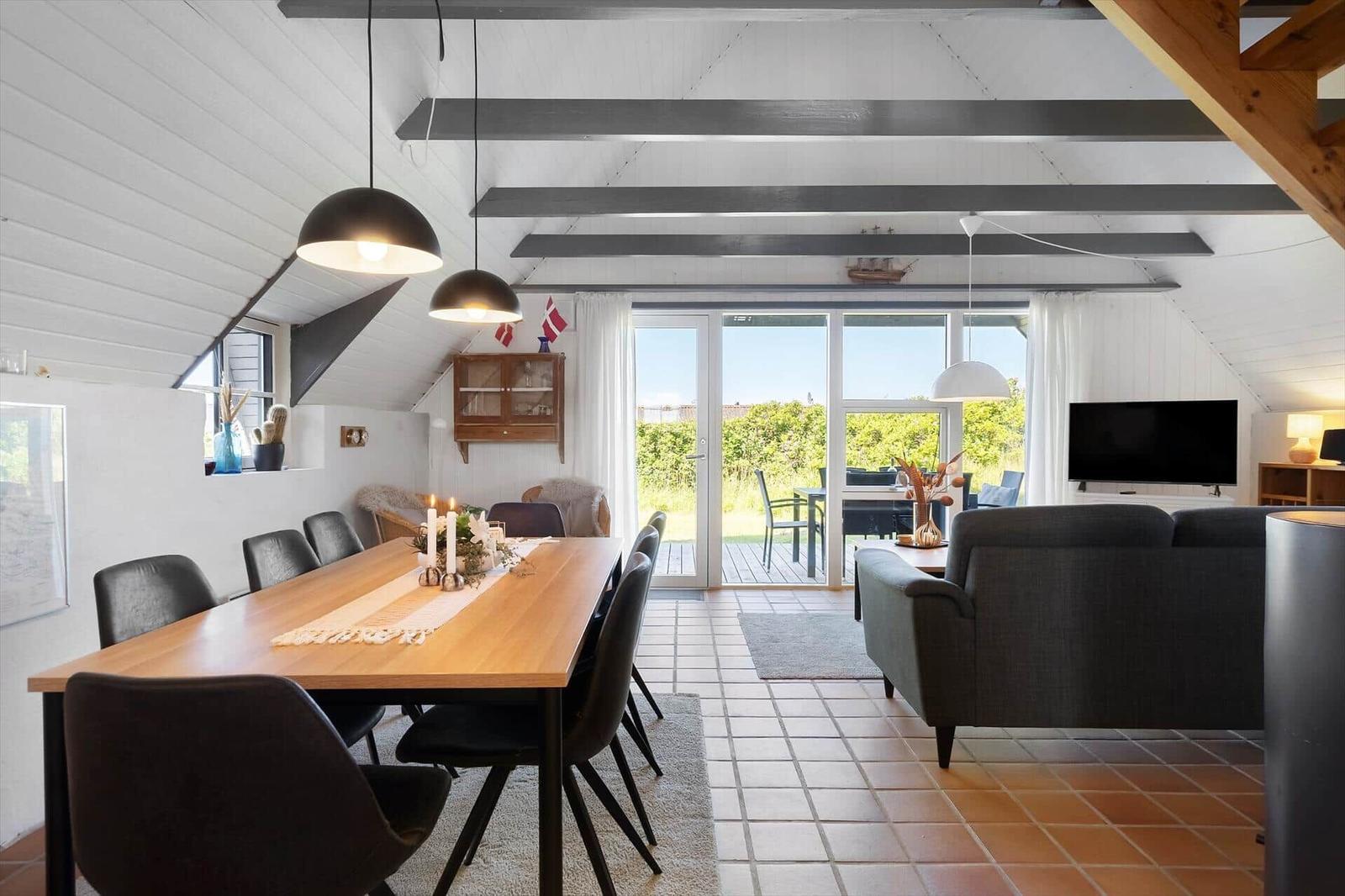 Wohn- und Essbereich mit Holztisch, Sofa und Terrassentür. Deckenbalken und Terrasse sind sichtbar.