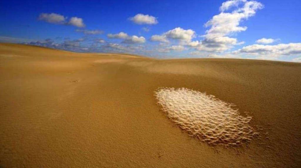 Wüstenlandschaft mit Sanddünen und einem weißen Sandbereich unter blauem Himmel mit Wolken.