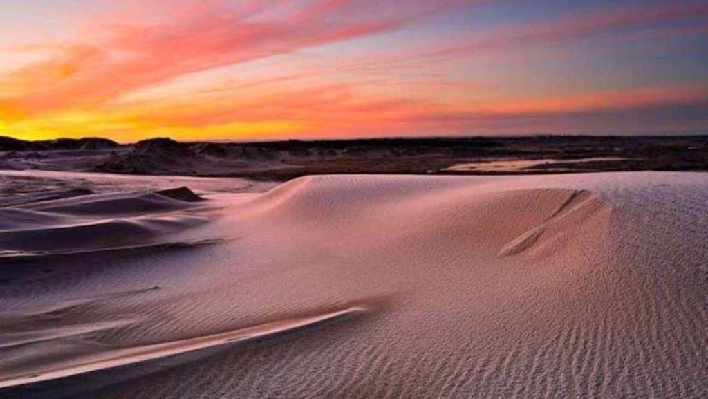 Weite Sanddünen unter einem farbenfrohen Sonnenuntergang am Horizont.