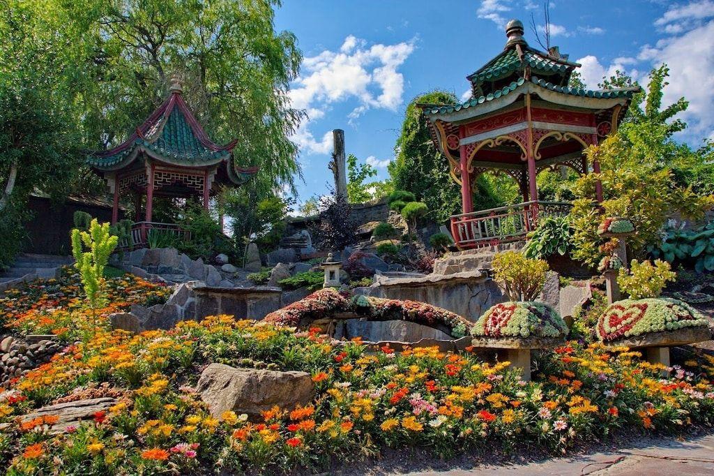 Zwei traditionelle Pavillons stehen in einem blühenden Garten mit bunten Blumen und Steinstrukturen.