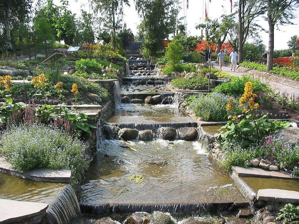 Ein Garten mit Wasserfall, Steinen und bunten Blumen. Menschen spazieren auf einem Weg.