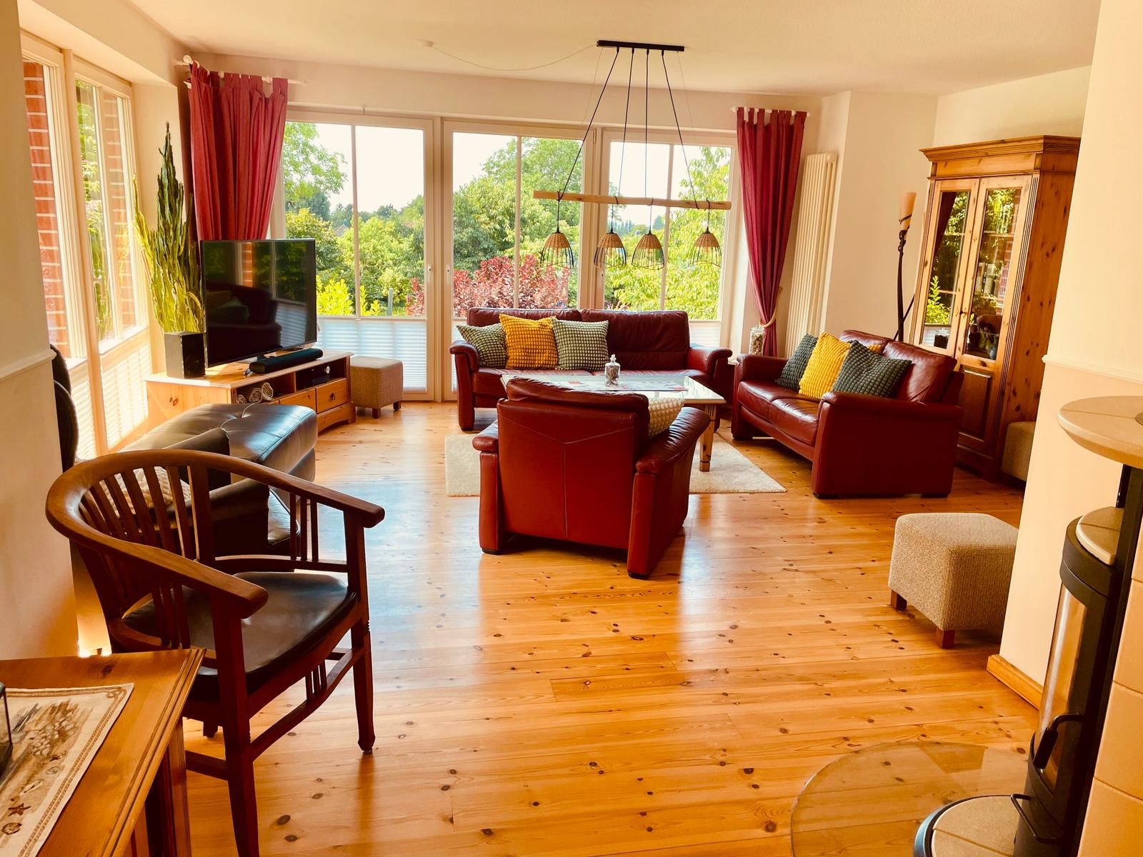 Wohnzimmer mit Holzboden, roten Ledersofas und großen Fenstern mit Blick auf Garten.