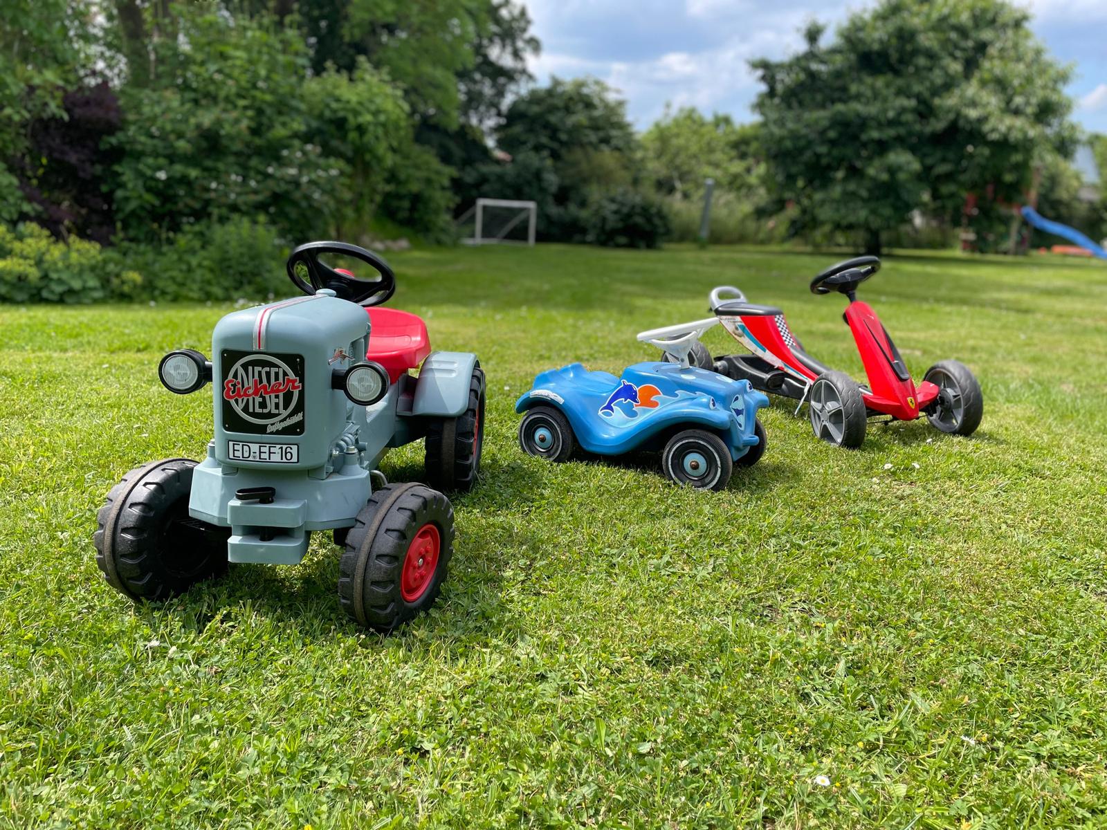 Drei Kinderfahrzeuge stehen auf dem Rasen. Ein Traktor, ein blaues Auto und ein rotes Karts.