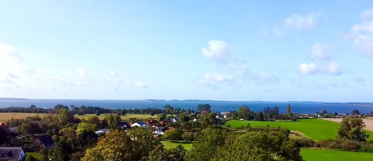 Weitläufiger Blick auf Felder, Häuser und einen See unter blauem Himmel mit vereinzelten Wolken.