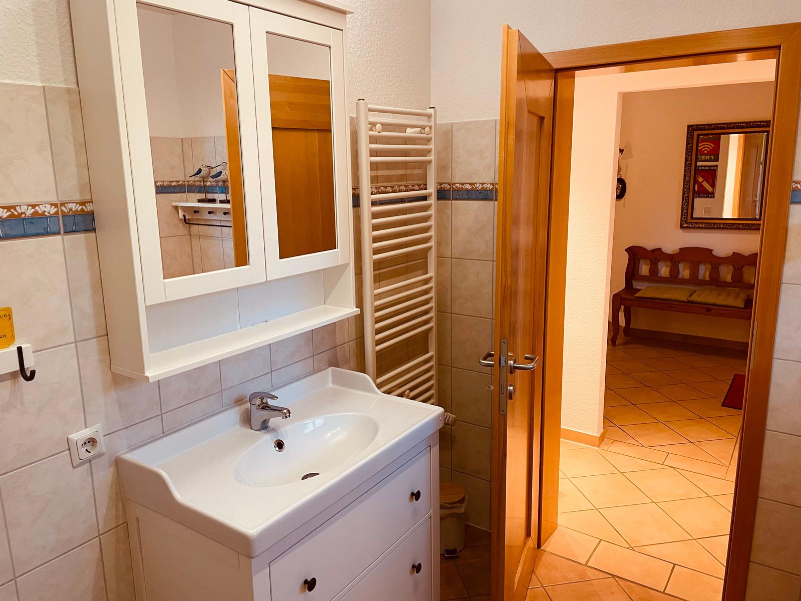 Badezimmer mit Waschbecken, Spiegelschrank und Heizkörper. Durchgang zu Wohnbereich mit Bank.