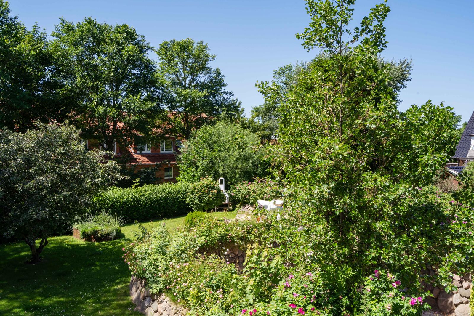 Garten mit Blumenbeeten, Grasfläche und Blick auf ein Haus hinter Bäumen.