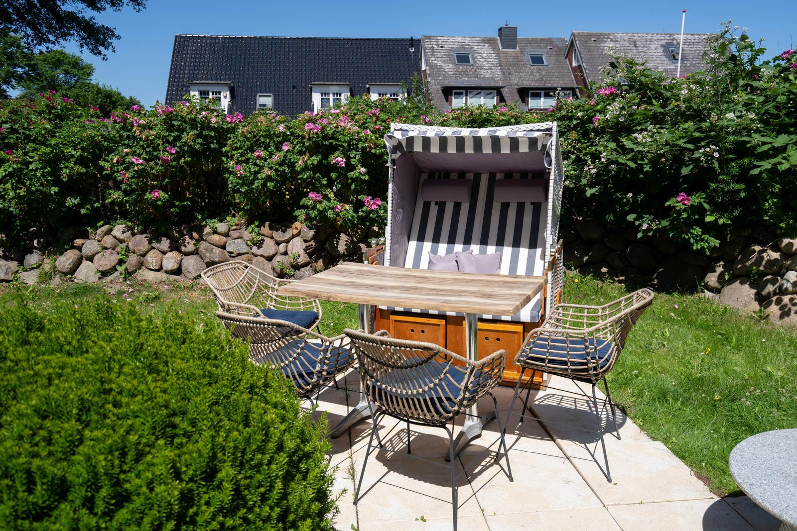 Hinterhof mit Strandkorb, Tisch und vier Stühlen. Rosenhecke und Steinmauer im Hintergrund.
