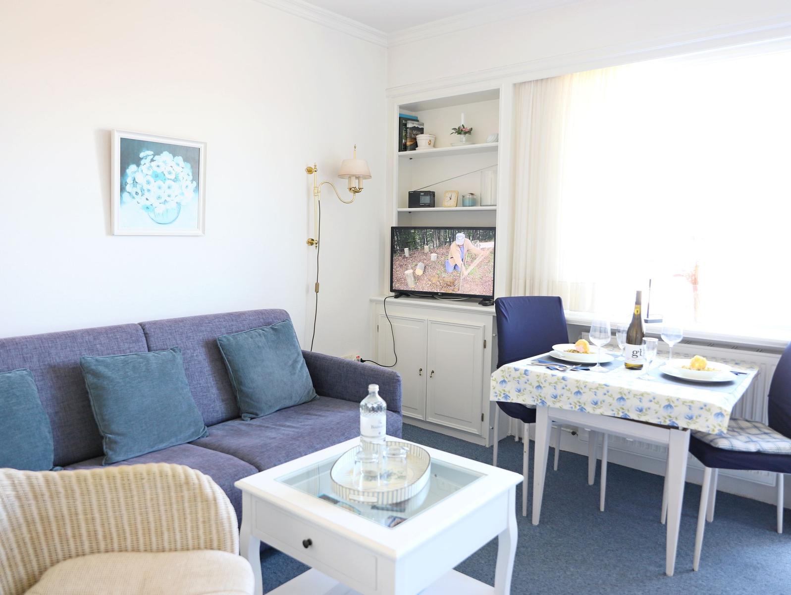 Wohnzimmer mit Sofa, Esstisch und Fernseher. Fenster mit Vorhängen. Tisch mit Tischdecke und Flasche.