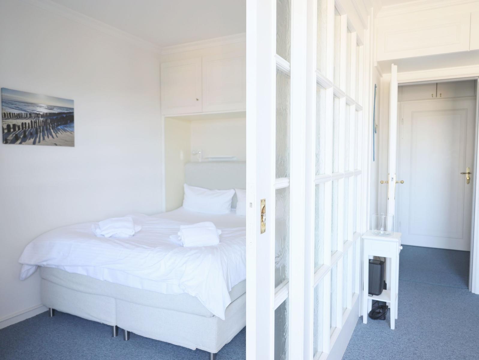 Schlafzimmer mit Bett, Wandbild und Tür. Blauer Teppich und weiße Möbel.