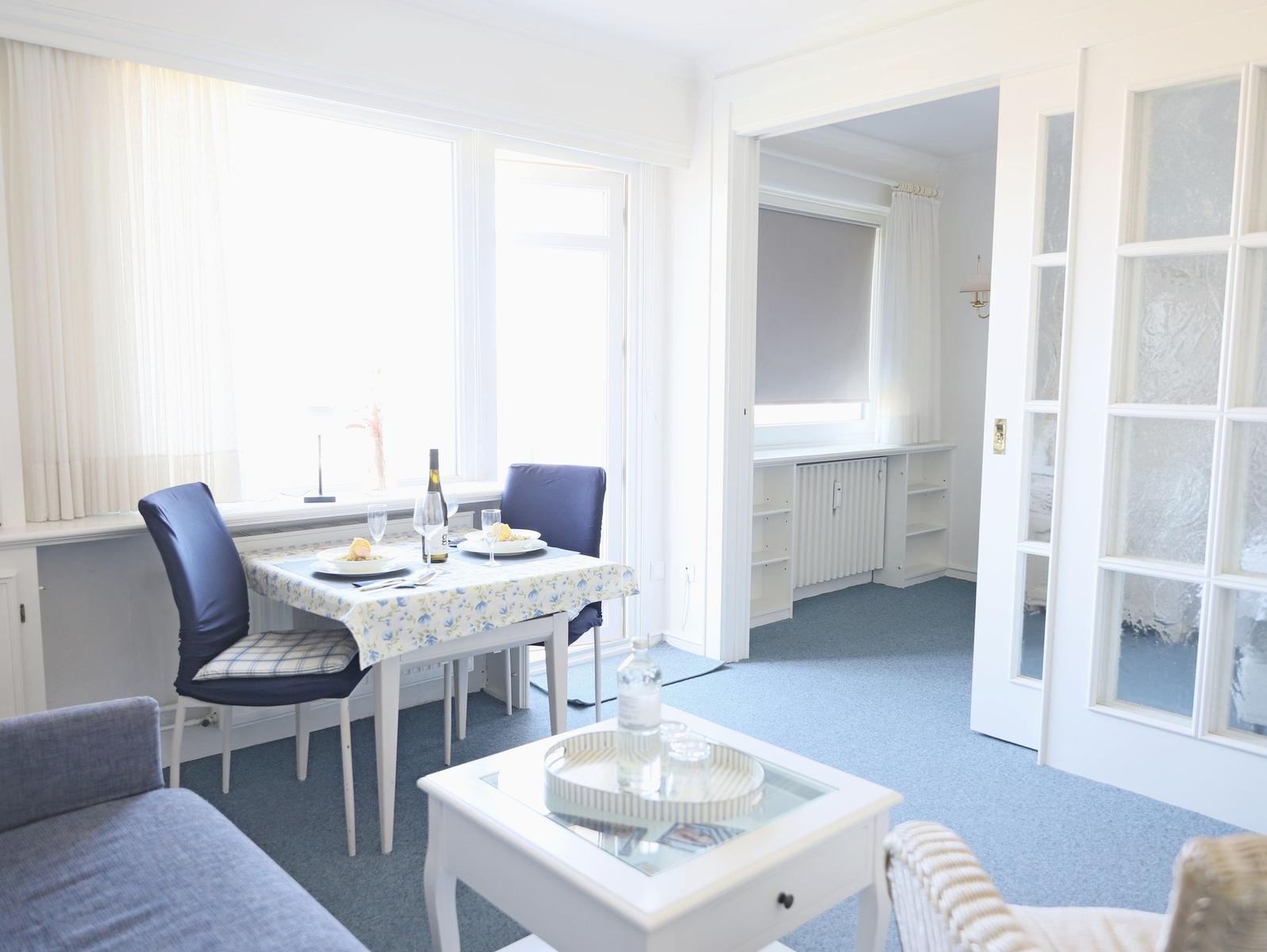Esszimmer mit Tisch, Stühlen und Fenster. Durchgang zu einem Raum mit Regalen.