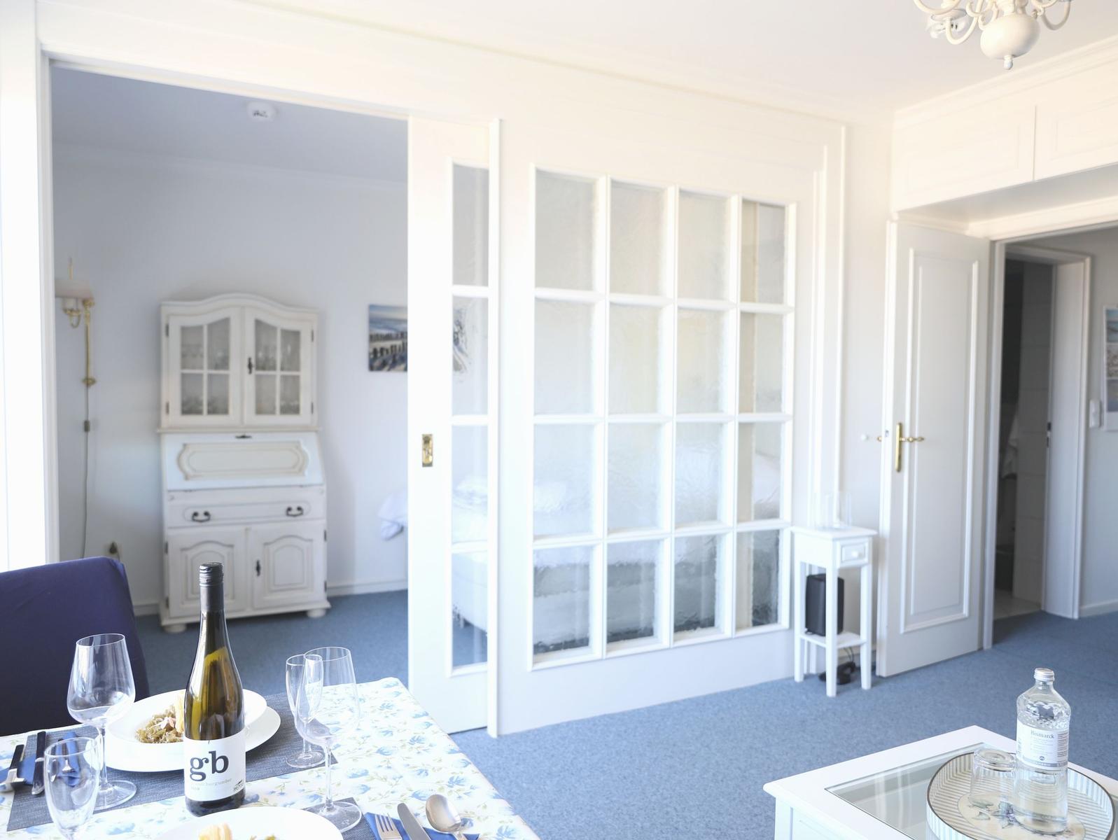 Essbereich mit Tisch, Flasche und Gläsern. Durchgehendes Fenster mit Glaspartien. Hintergrund: Schrank und Tür.