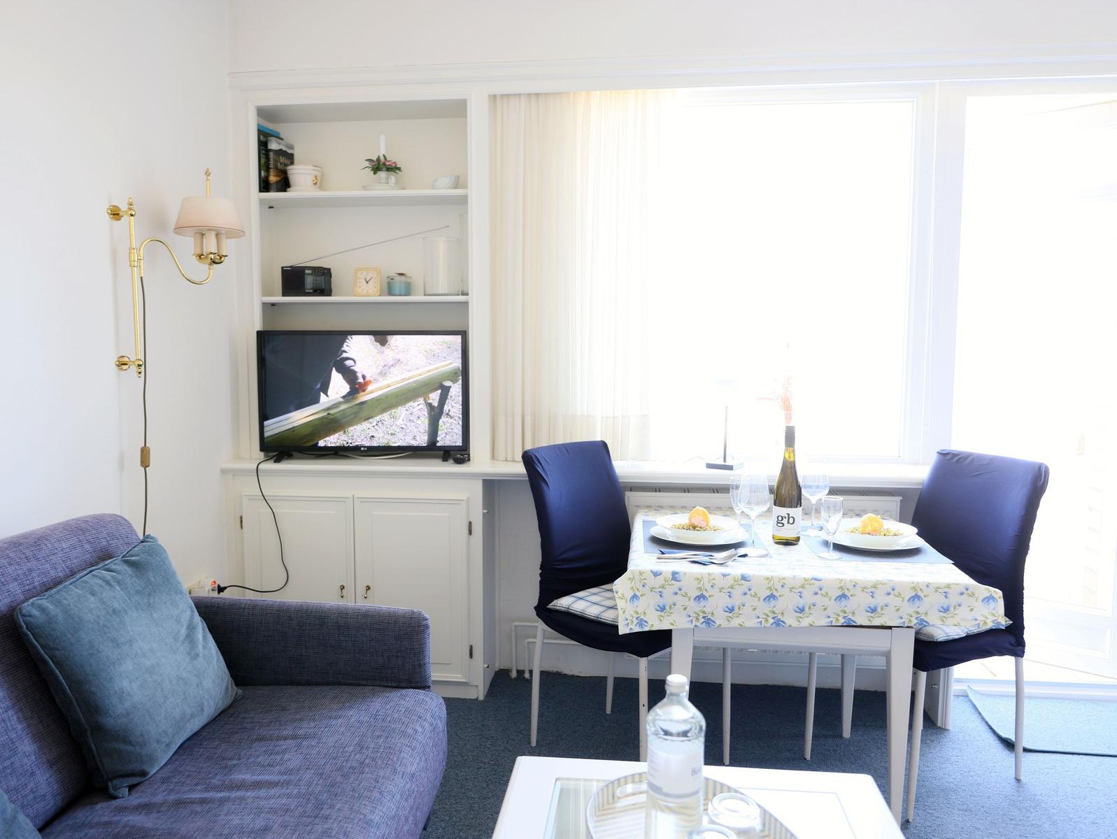 Wohnzimmer mit Sofa, Esstisch und Fernseher. Fenster mit Vorhängen und Tisch mit Speisen.