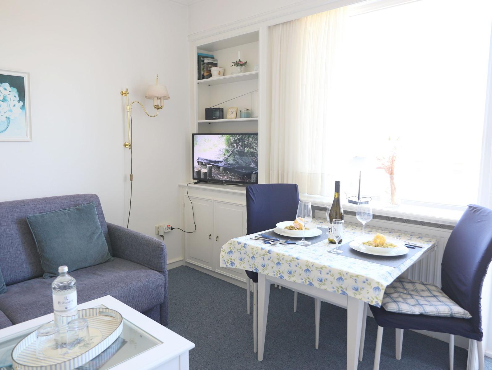 Wohn- und Essbereich mit Sofa, Tisch und TV. Fenster mit Vorhängen.
