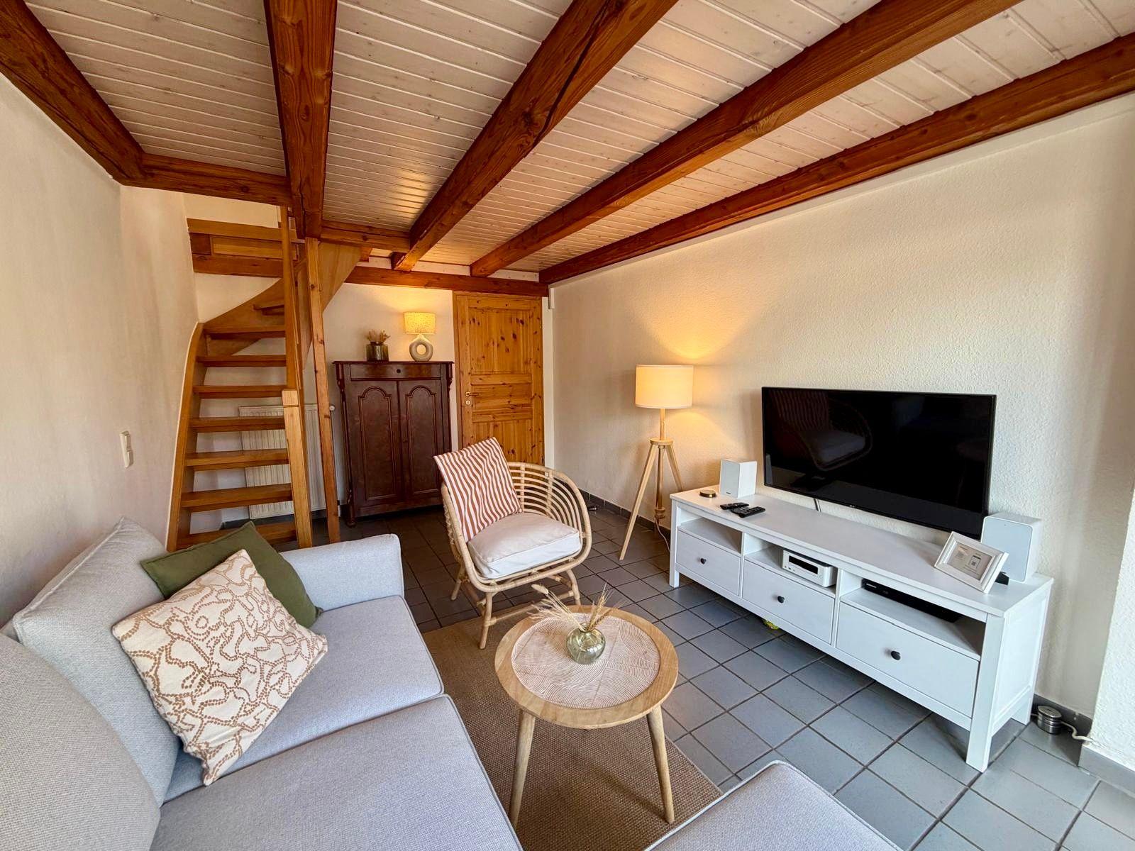 Wohnzimmer mit Sofa, Fernseher und Holzbalkendecke. Treppe führt zur oberen Etage.