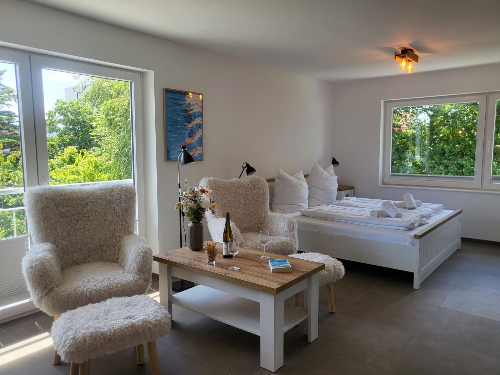 Ein Schlafzimmer mit Bett, zwei Flauschstühlen und Tisch. Fenster bieten Blick auf Grün.