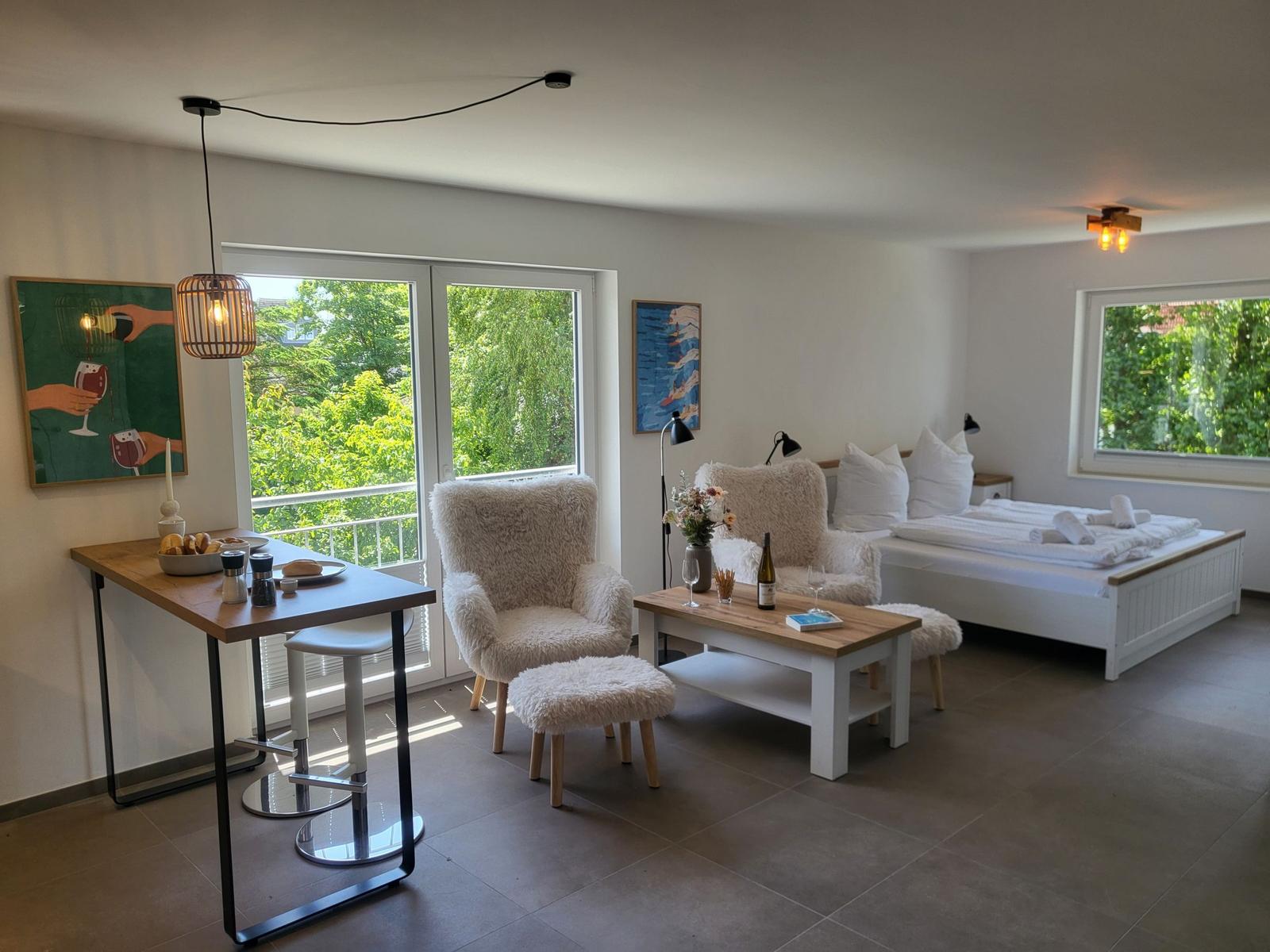 Ein Raum mit Schlafplatz, Sitzgelegenheiten und Tisch. Durchgänge führen zu Balkon und Fenster.