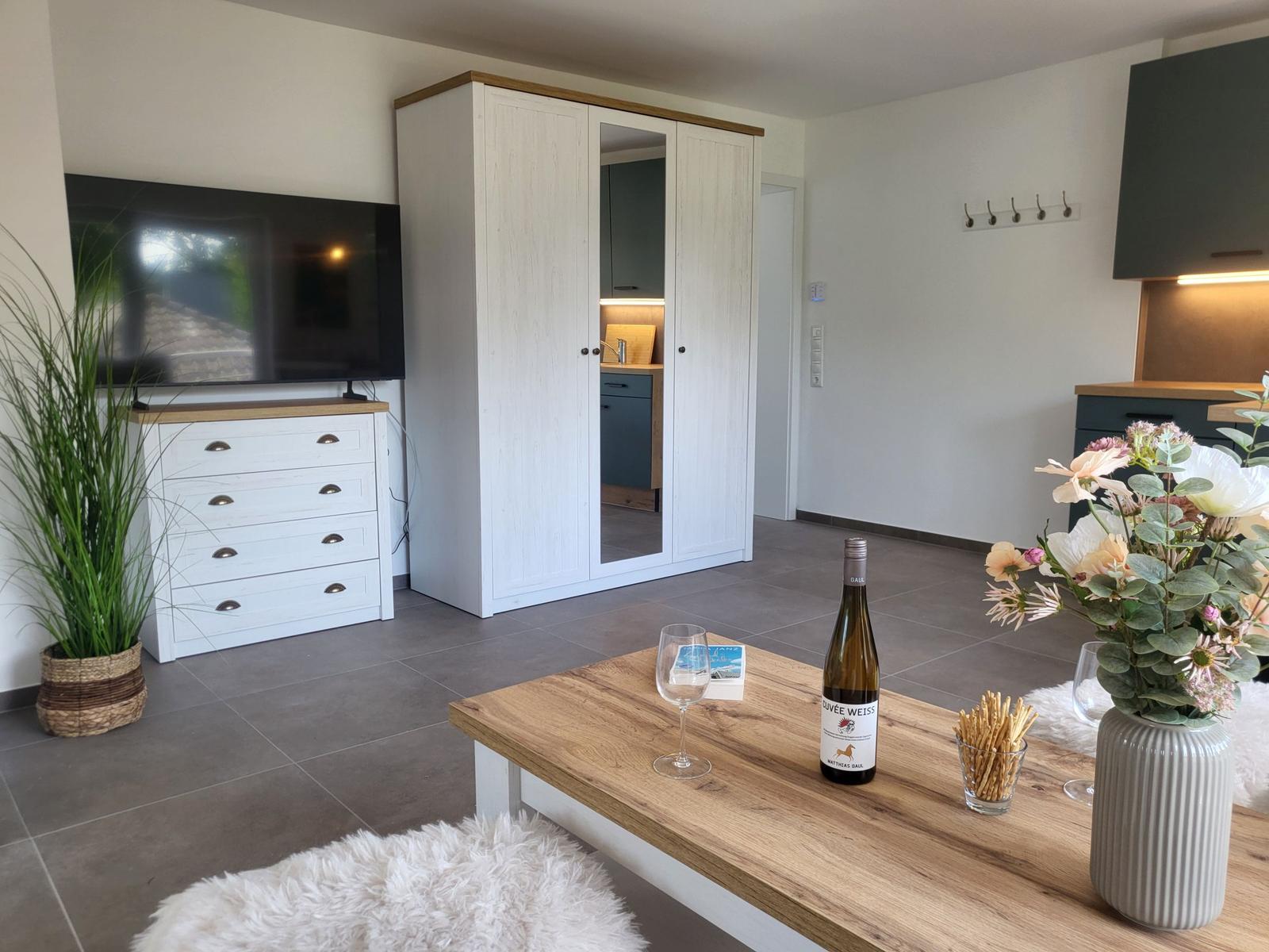 Wohnbereich mit TV, Schrank und Tisch mit Wein und Blumen.
