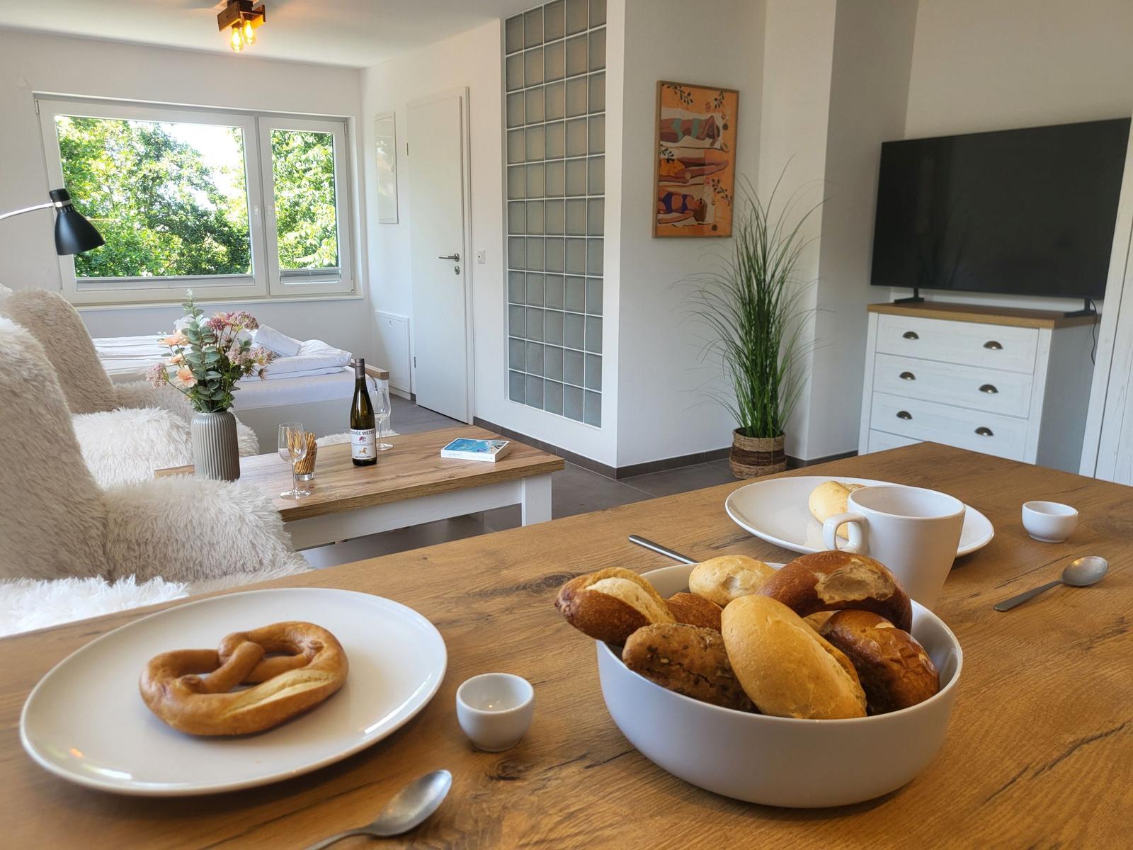 Esstisch mit Brot und Kaffee, Wohnbereich mit Fernseher und Fenster.