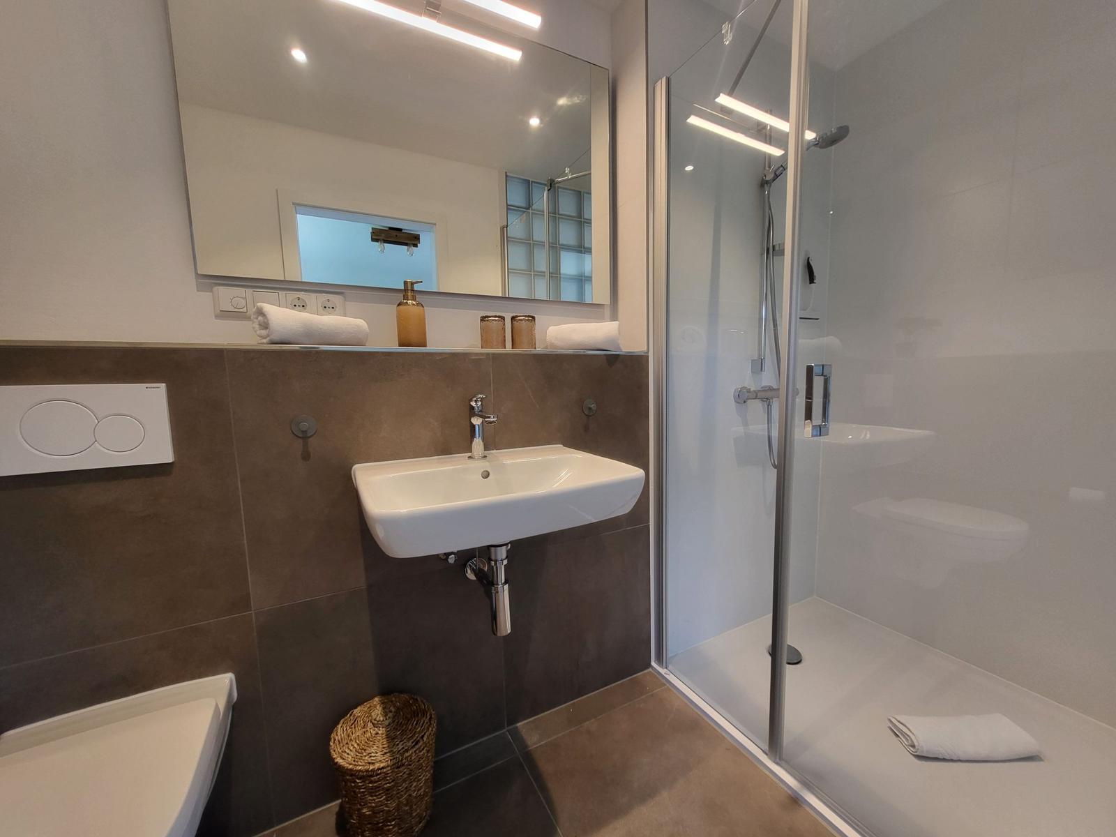 Badezimmer mit Dusche, Waschbecken und Spiegel. Toilette und Handtuch sind sichtbar.