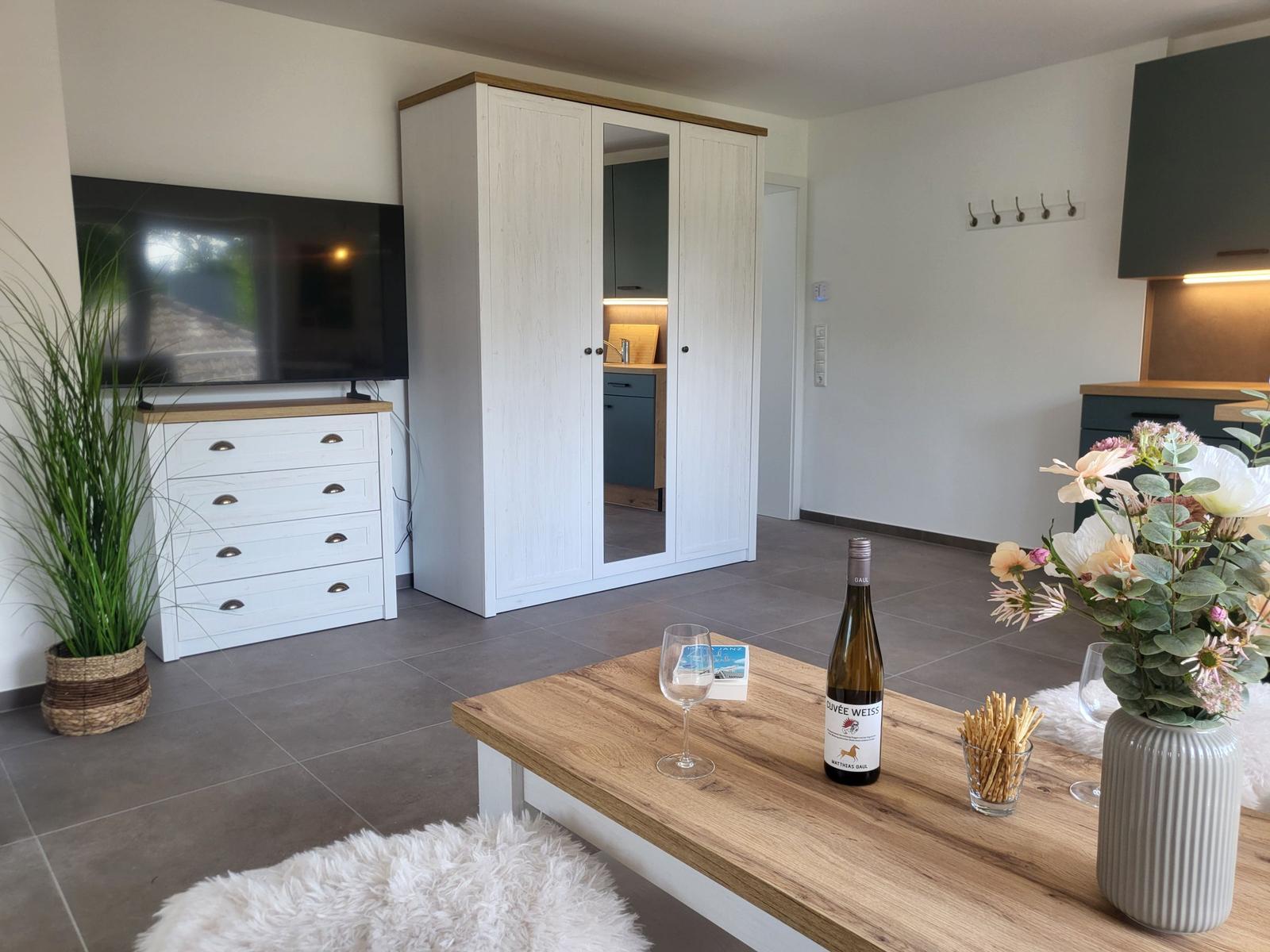Wohnbereich mit TV, Schrank und Tisch mit Wein und Blumen.
