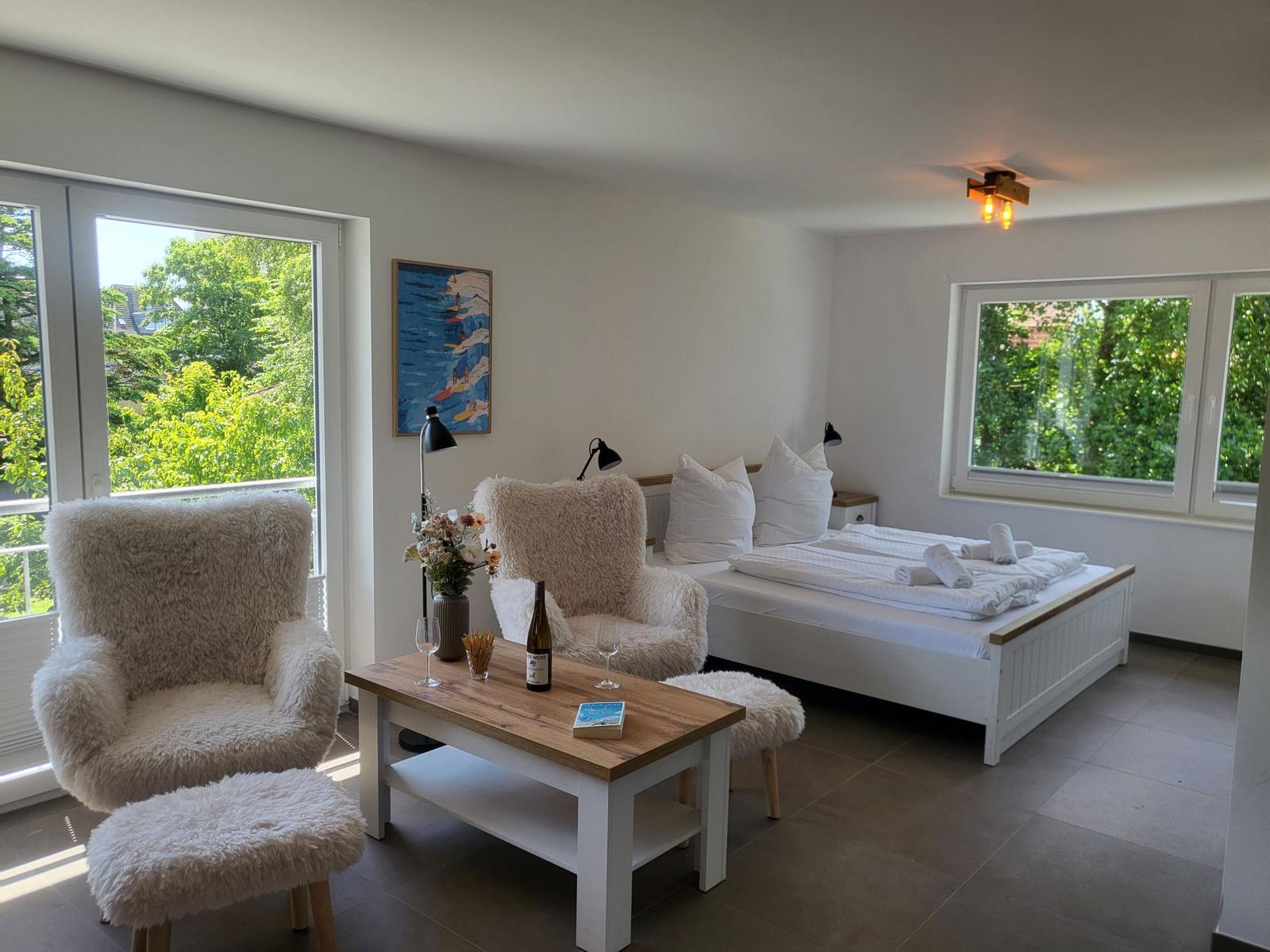 Ein Schlafzimmer mit Bett, zwei Stühlen und Tisch. Durch Fenster blickt man auf Grün.