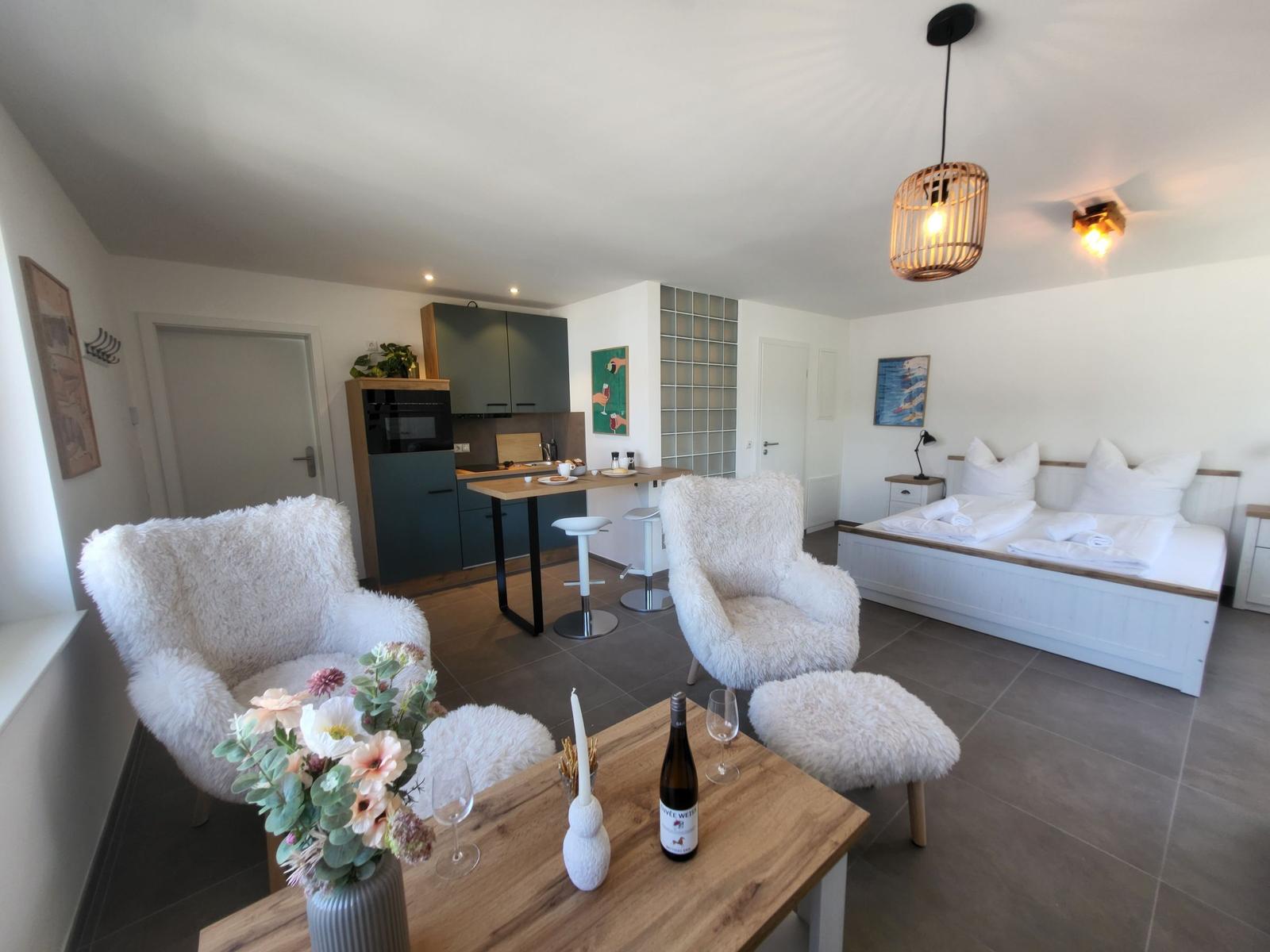 Wohnbereich mit Schlafzimmer, Küche und zwei weißen Sesseln. Tisch mit Blumen und Weinflasche.