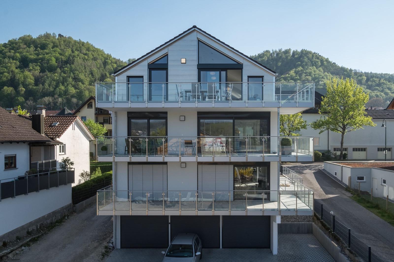 Moderne Villa mit drei Terrassen und großem Balkon. Garagenbereich und grüne Hügel im Hintergrund.