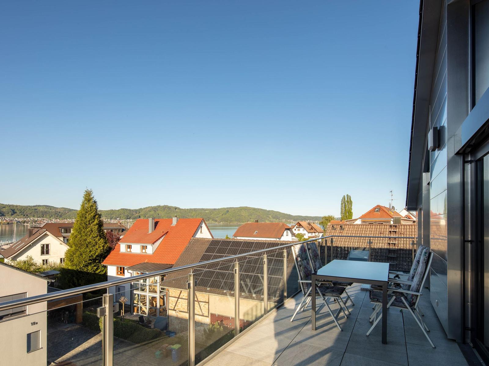 Terrasse mit Tisch und Stühlen, Blick auf Dächer und See unter blauem Himmel.