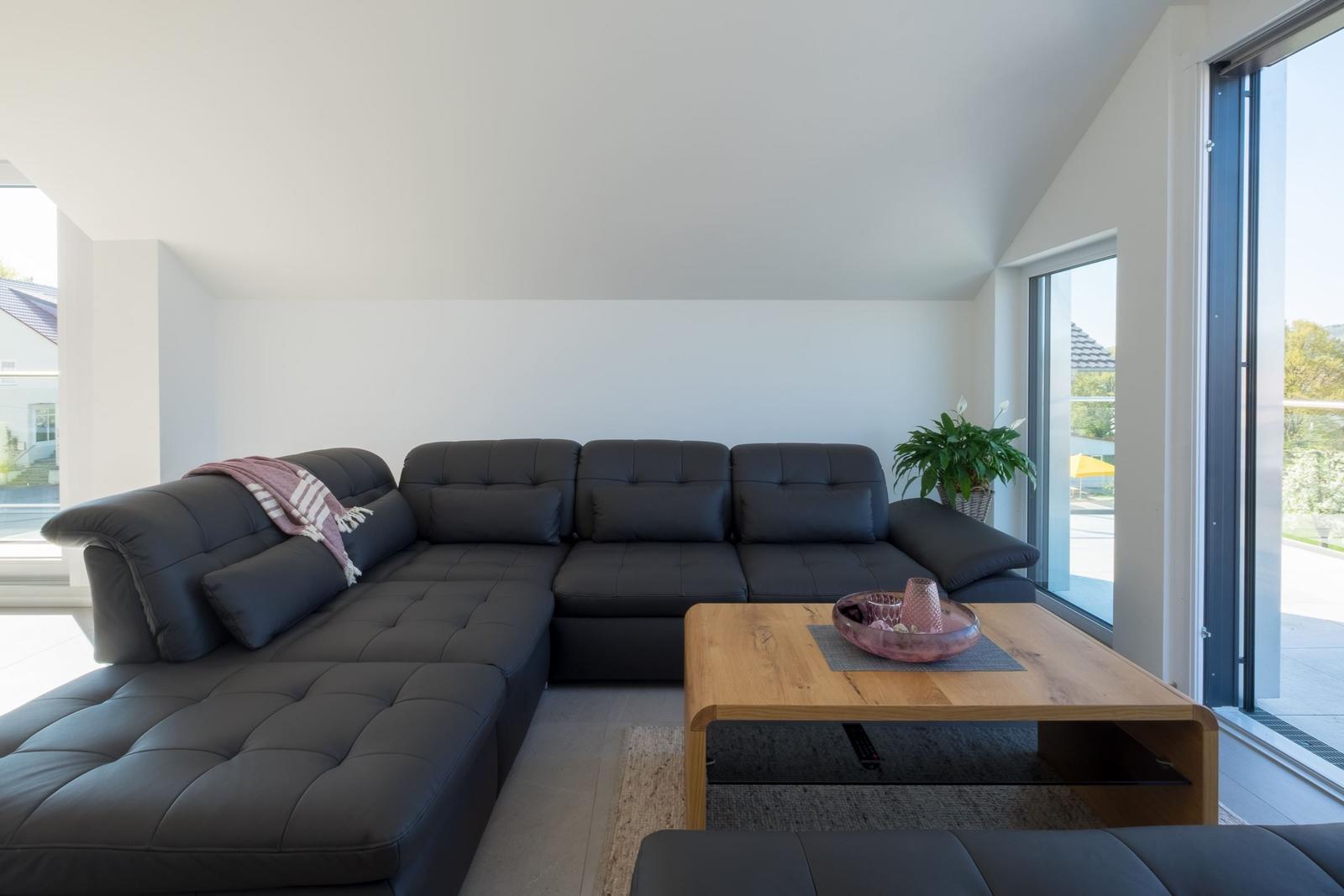 Ein Wohnzimmer mit einem schwarzen Sofa, einem Holztisch und großen Fenstern.