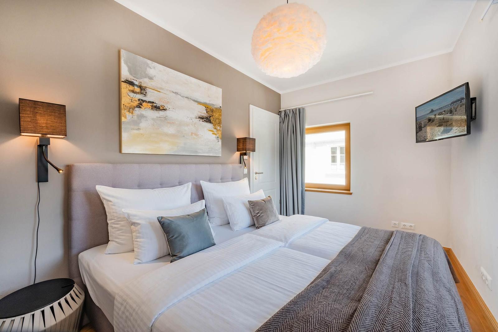 Schlafzimmer mit Bett, Wandbild, Deckenleuchte und TV. Fenster mit Vorhängen.