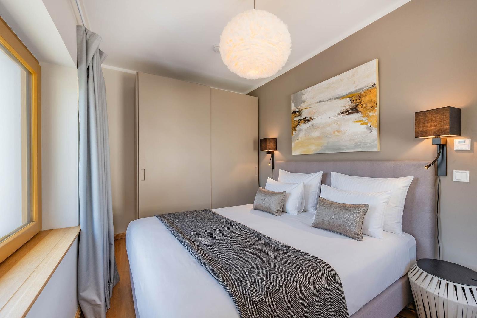 Schlafzimmer mit Bett, Wandbild und Deckenleuchte. Fenster mit Vorhang und Nachttisch.